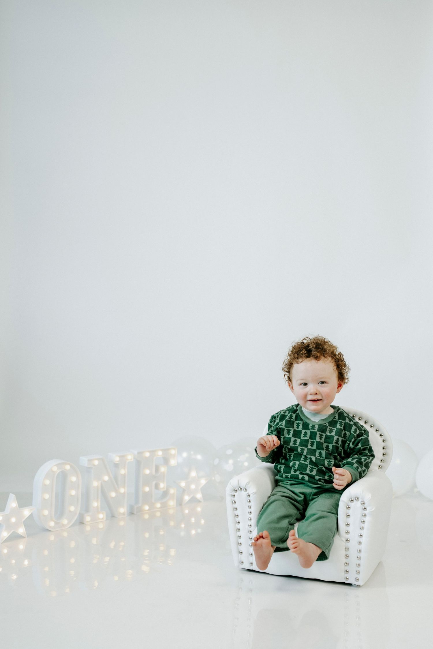 A small child in a green outfit sits on a white cushion with glowing letter lights in the background.