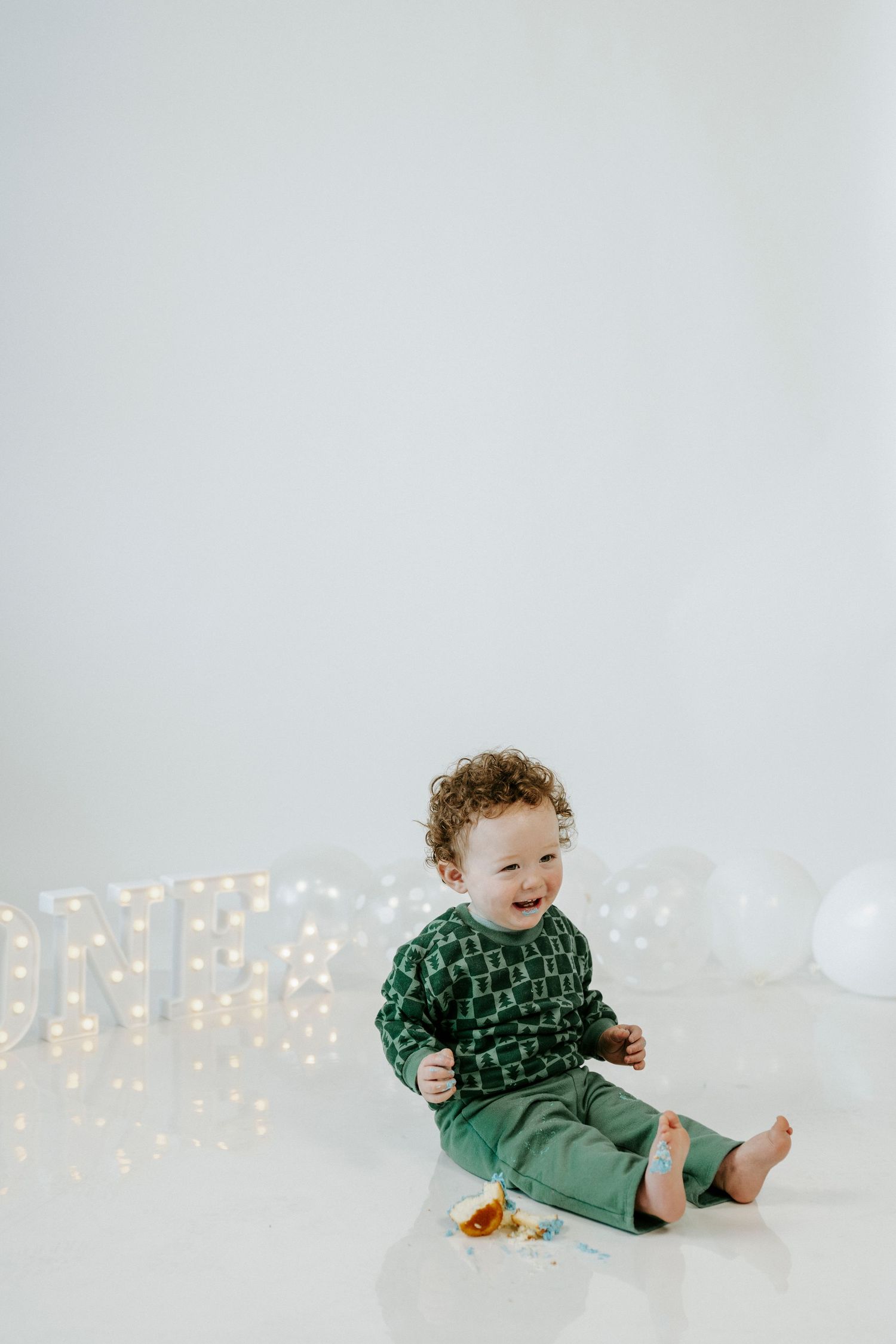 Baby in green dress sits on white floor with twinkling fairy lights in background creating magical holiday atmosphere.