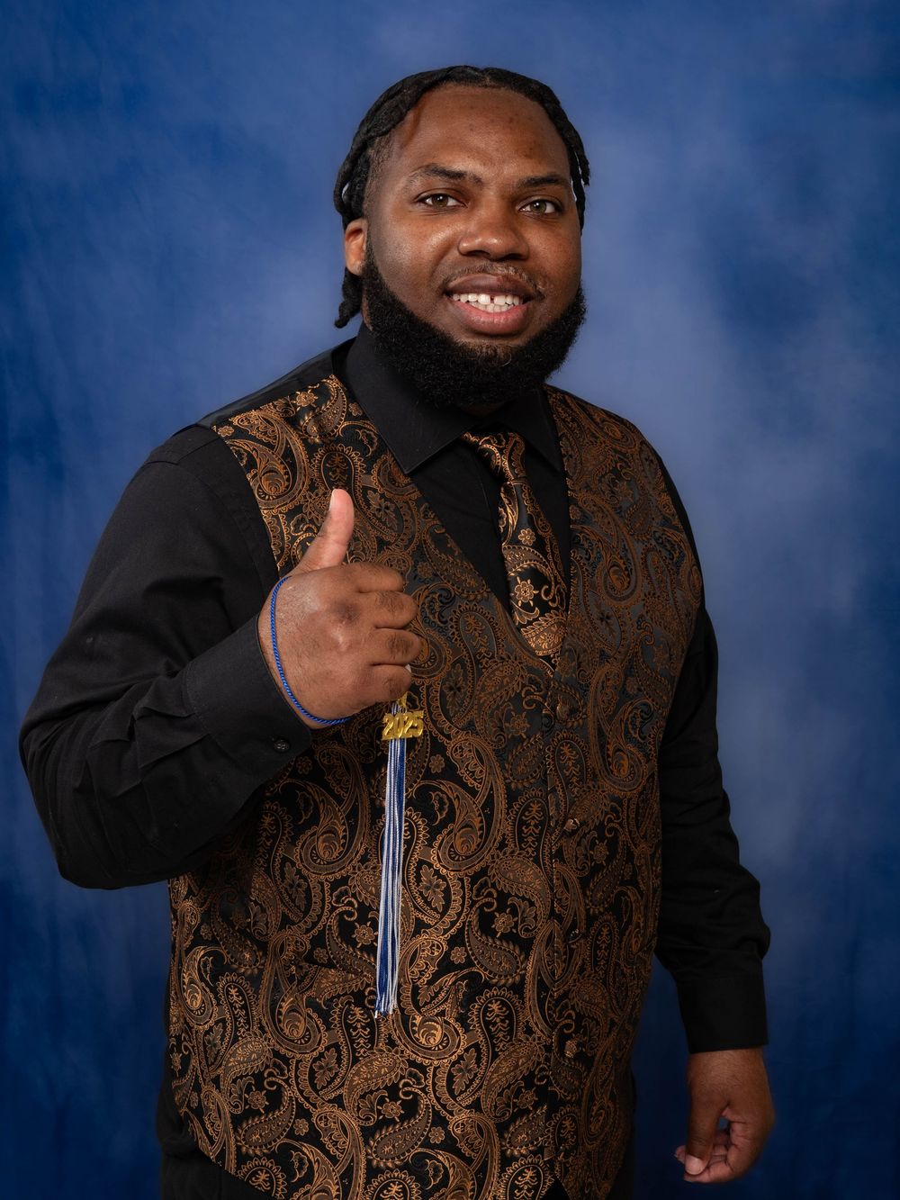 Professional portrait of someone in a black dress shirt and brown patterned vest giving a thumbs up against a blue backdrop.