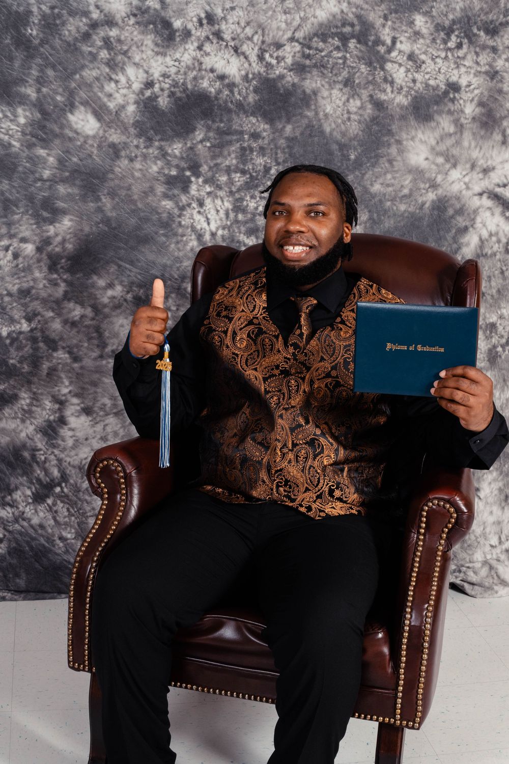Graduate celebrating academic achievement with diploma while sitting in elegant leather armchair against gray backdrop.