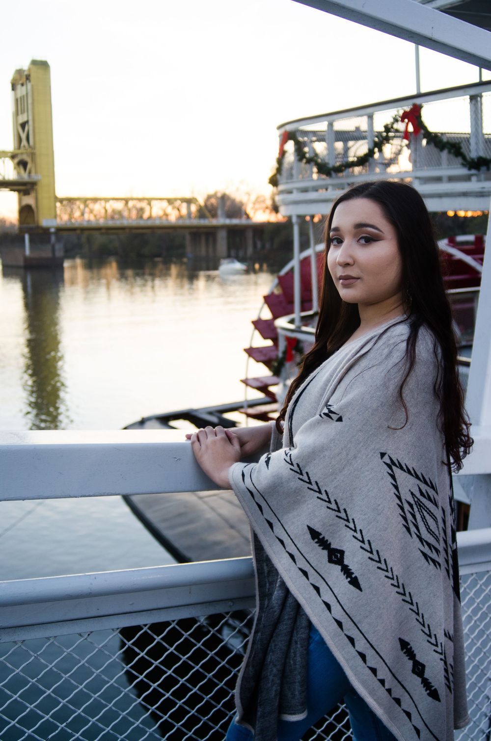 A person in a gray sweater leans against a railing at sunset near a waterfront bridge.