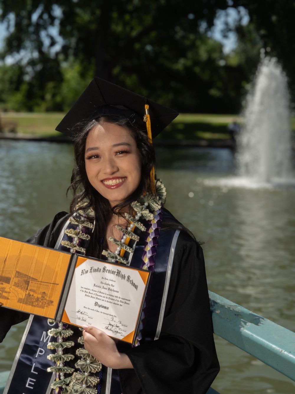 Graduate in cap and gown poses with diploma and fountain backdrop in outdoor graduation photoshoot.