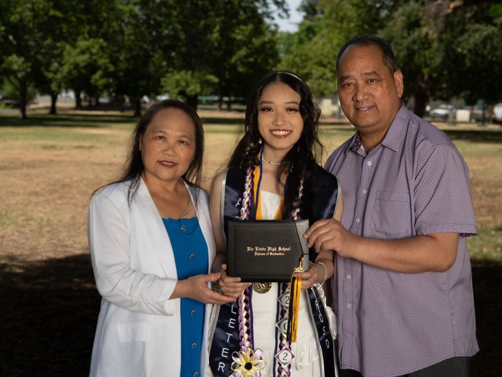 Three people pose outdoors with a graduation diploma in a park setting with trees in the background.