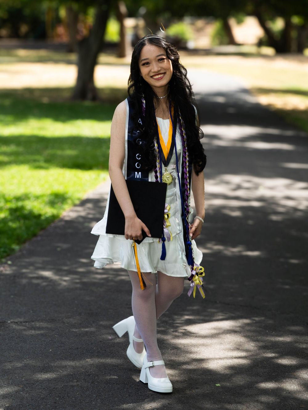 A graduate in a black vest and white dress poses with a diploma while standing on a tree-lined path on graduation day.