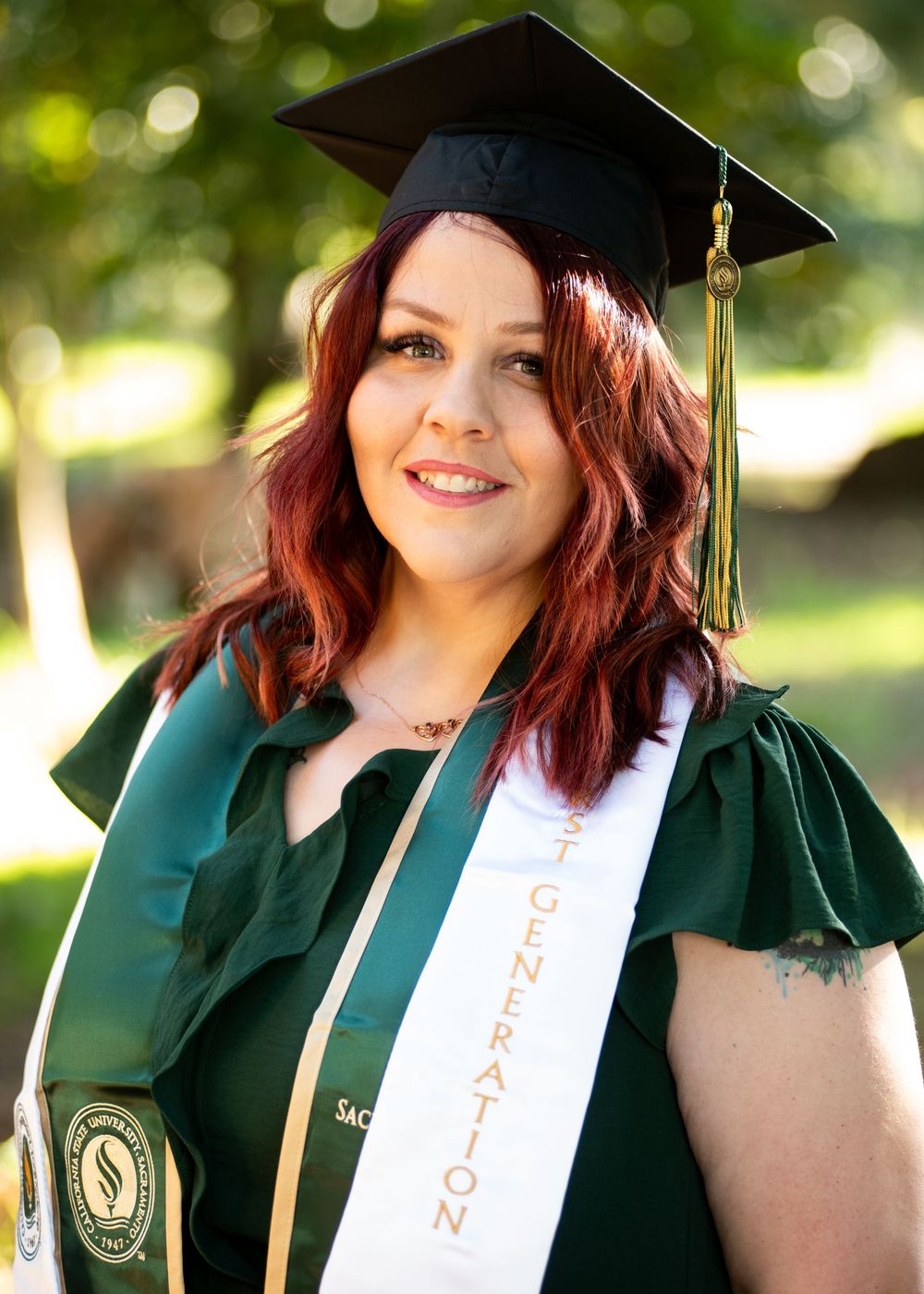 Graduate in dark green cap and gown with white sash poses outdoors with natural bokeh background.