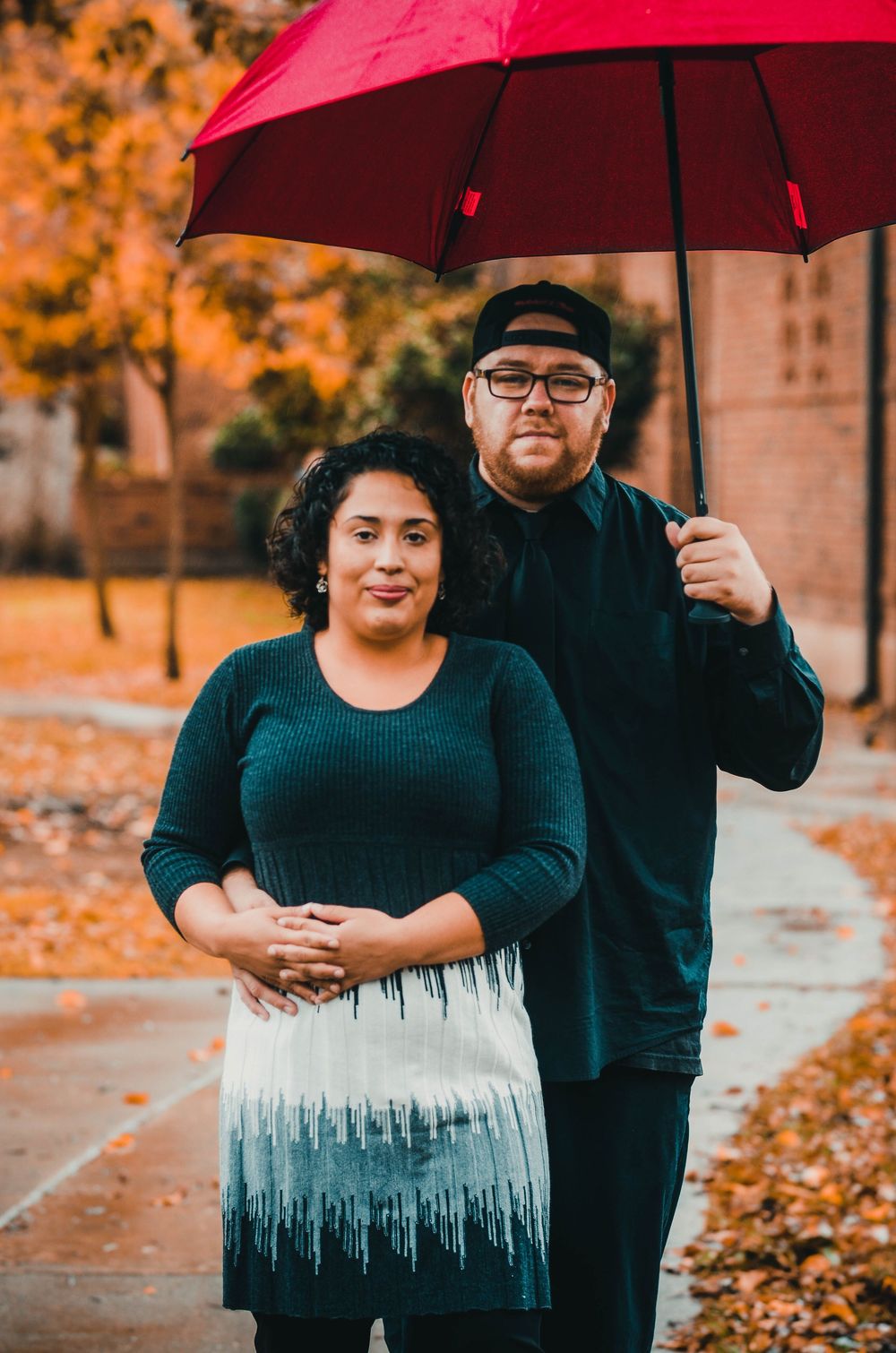 A couple stands together under a red umbrella on an autumn day with fallen leaves and brick buildings in the background.