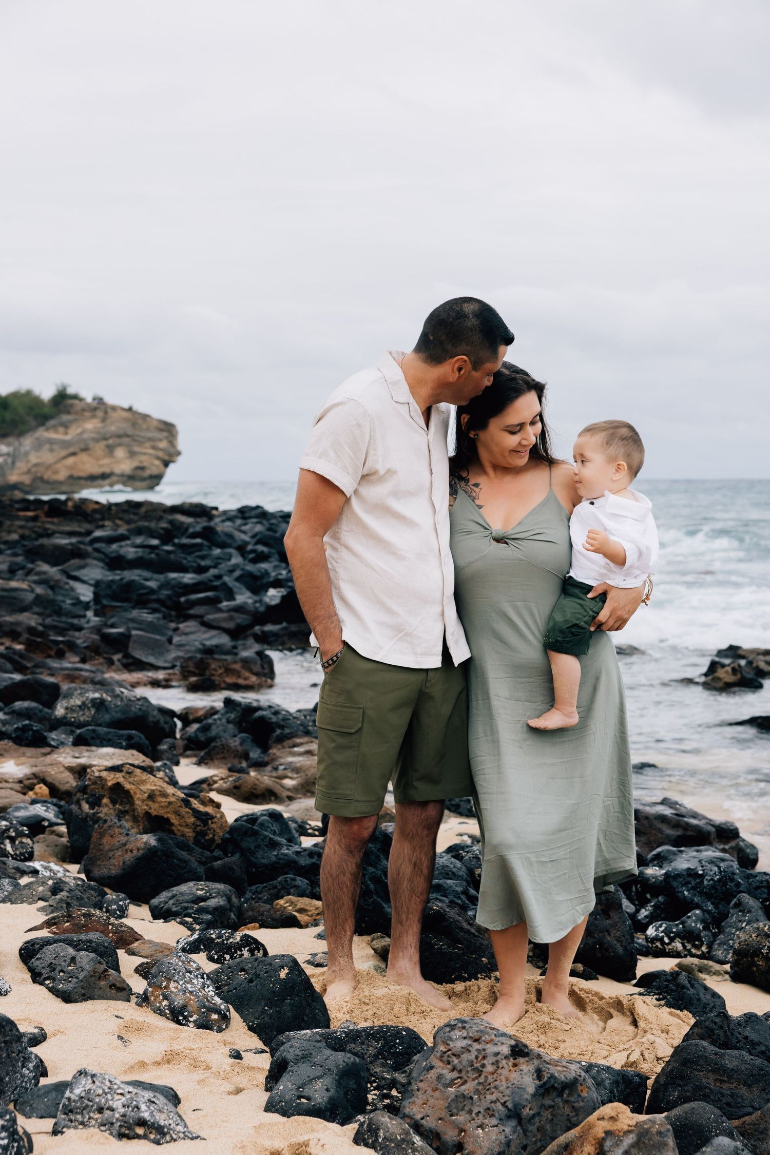 Young family of three standing on rocky Hawaiian beach shore on cloudy day.