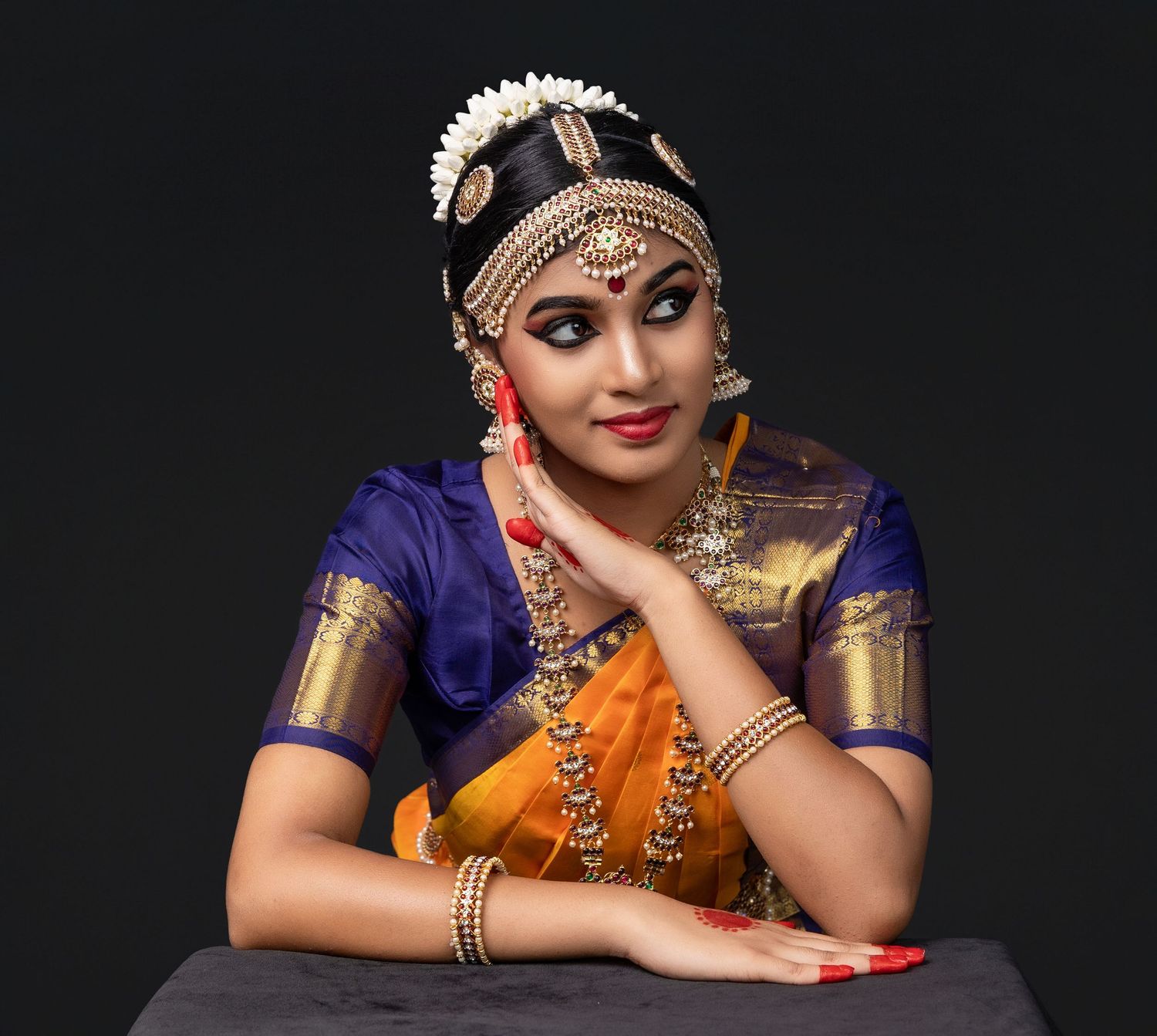 Traditional Indian bride in royal blue and orange saree with gold jewelry poses elegantly against dark background.