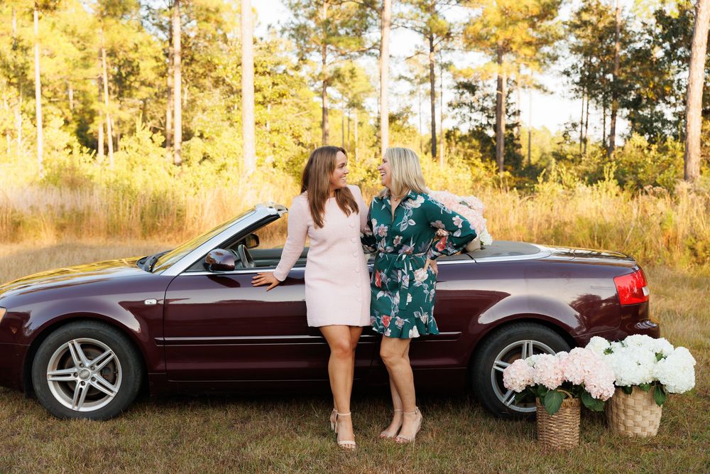 Two people stand in a golden autumn forest near a maroon convertible car during a sunset photo session.