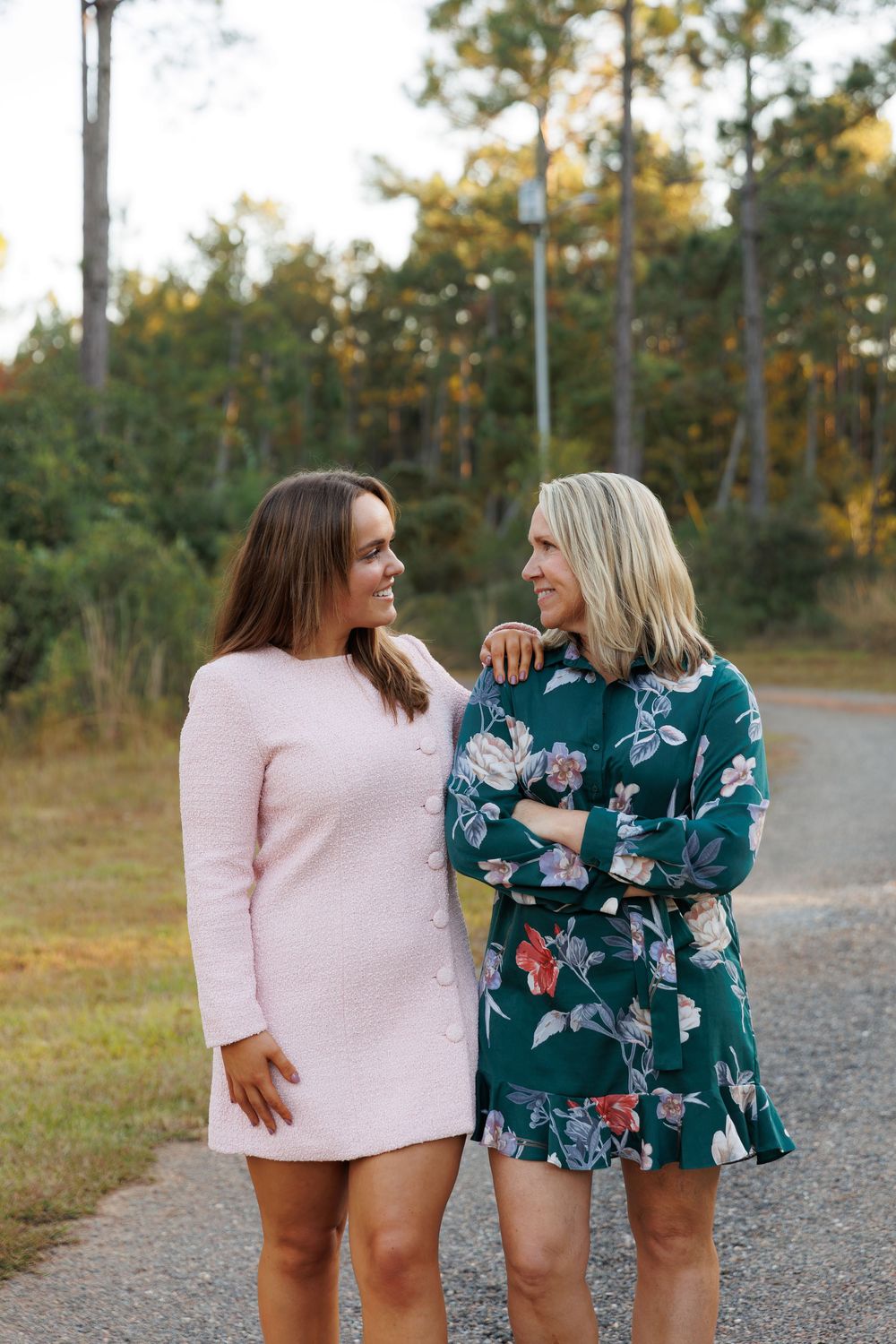 Two women in contrasting pink and floral outfits laugh together on a gravel path with pine trees in the background.