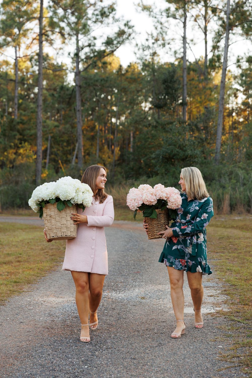Two friends carrying large floral arrangements in woven baskets while walking on a path through pine trees at sunset.