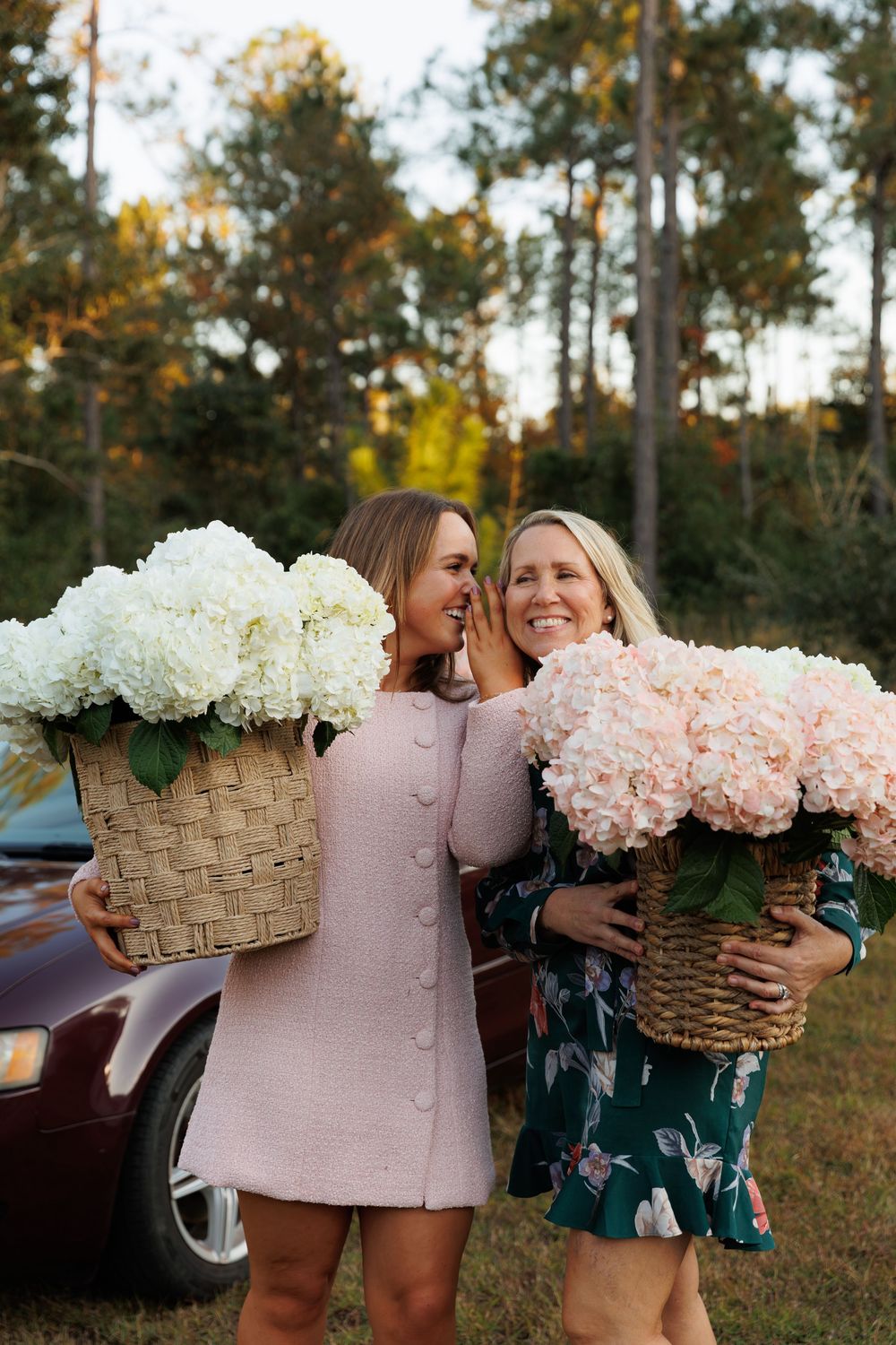 Two friends laughing together while holding beautiful hydrangea and rose flower arrangements in woven baskets outdoors.