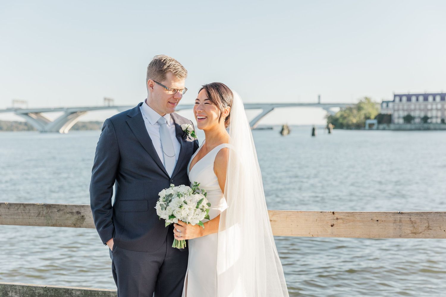 A couple in wedding attire standing by a waterfront with a bridge visible in the distance.