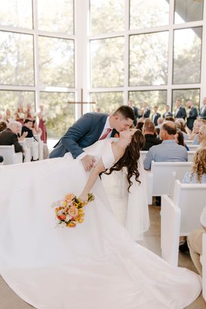 Wedding guests performing synchronized dance move in bright reception hall with large windows.