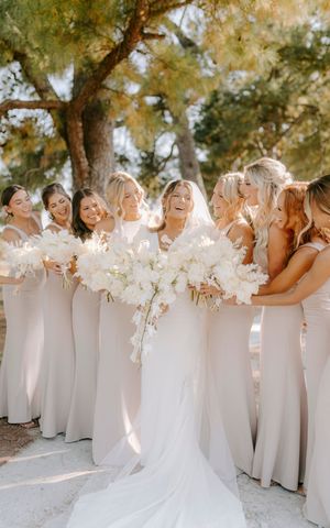 A joyful wedding party wearing white dresses holds white floral bouquets while standing together outdoors.