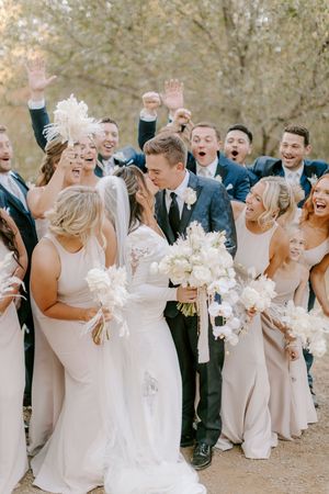 Wedding party in light colored dresses and dark suits share a joyful candid moment outdoors with white bouquets.