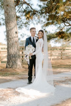 A romantic wedding couple embraces under tall pine trees in an outdoor setting with soft natural lighting.