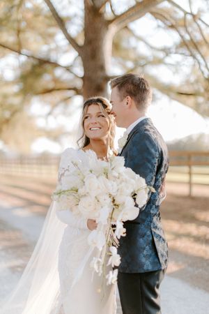 A couple shares a tender moment in a sunlit outdoor setting with white floral bouquet and elegant wedding attire.