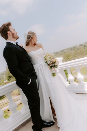 Bride and groom stand on elegant white balcony, she holds floral bouquet in wedding dress, he wears black tuxedo.