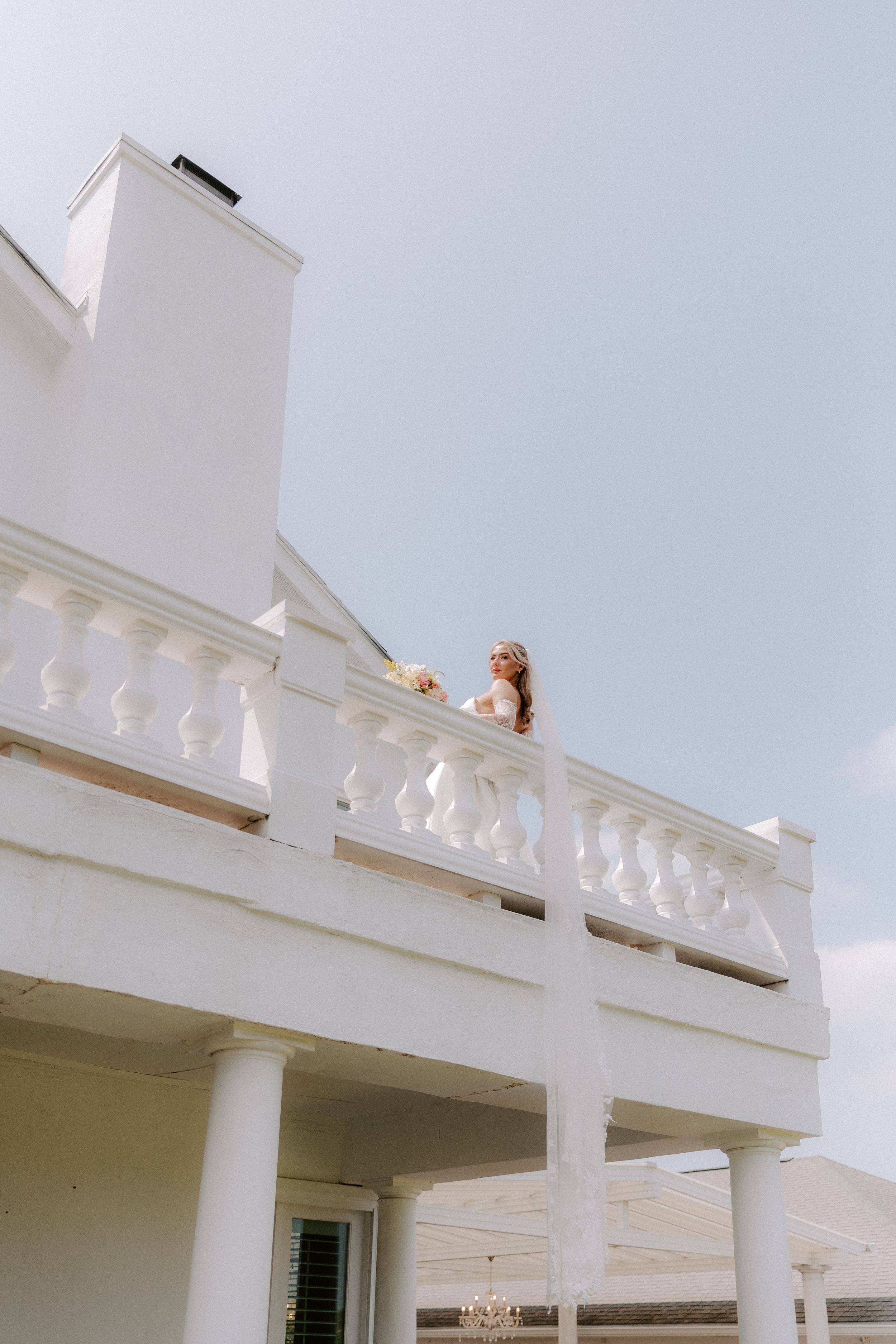 Woman in white dress holding a dog on elegant white balcony with classical columns against blue sky.