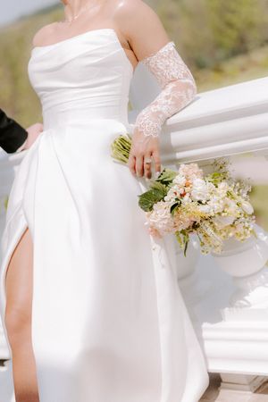 Bride in strapless white gown with lace gloves holding a pastel bouquet of pink and white flowers at an outdoor wedding.