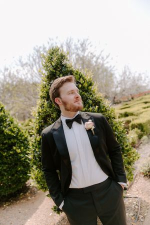 Groom in black tuxedo with bow tie and boutonniere stands confidently in a garden setting.