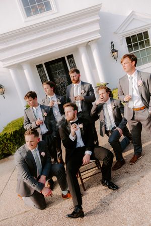 Groom and groomsmen in suits smoking cigars outside a white colonial venue during a wedding celebration.