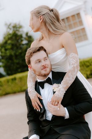 Bride in strapless gown with lace gloves embraces groom in black tuxedo outdoors at elegant wedding venue.