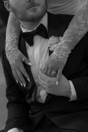 Black and white wedding photo of bride in lace gloves embracing groom in tuxedo with bow tie, showing rings.