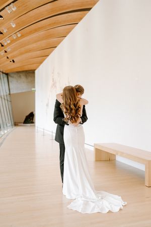 A couple in wedding attire shares an intimate moment in a bright, minimalist indoor space with wooden beams.