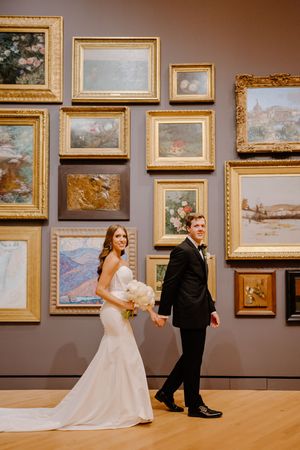 Two people in formal attire pose in front of a gallery wall displaying ornate gold-framed paintings in an art museum.