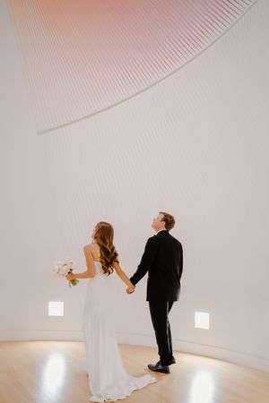 A couple in wedding attire dances together in an elegant white curved room with dramatic lighting.