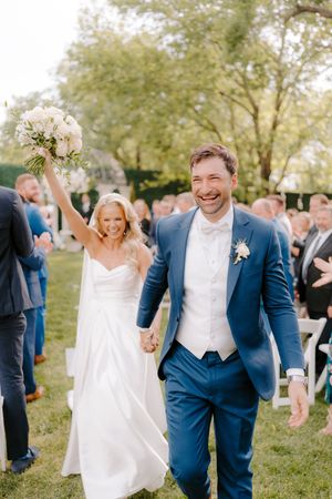 A joyful wedding couple walks down the aisle after their outdoor ceremony while guests celebrate around them.