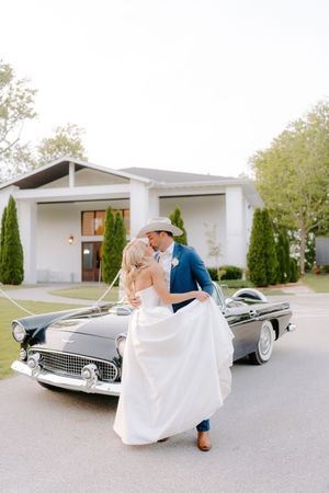 Romantic couple dancing beside a classic vintage car in front of a white building with elegant landscaping.