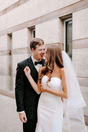 A romantic moment between newlyweds sharing laughter against a light stone building exterior.