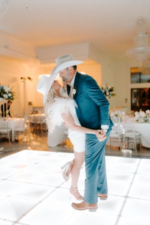 A couple shares their first dance at a wedding reception in an elegant white room with teal and romantic lighting.