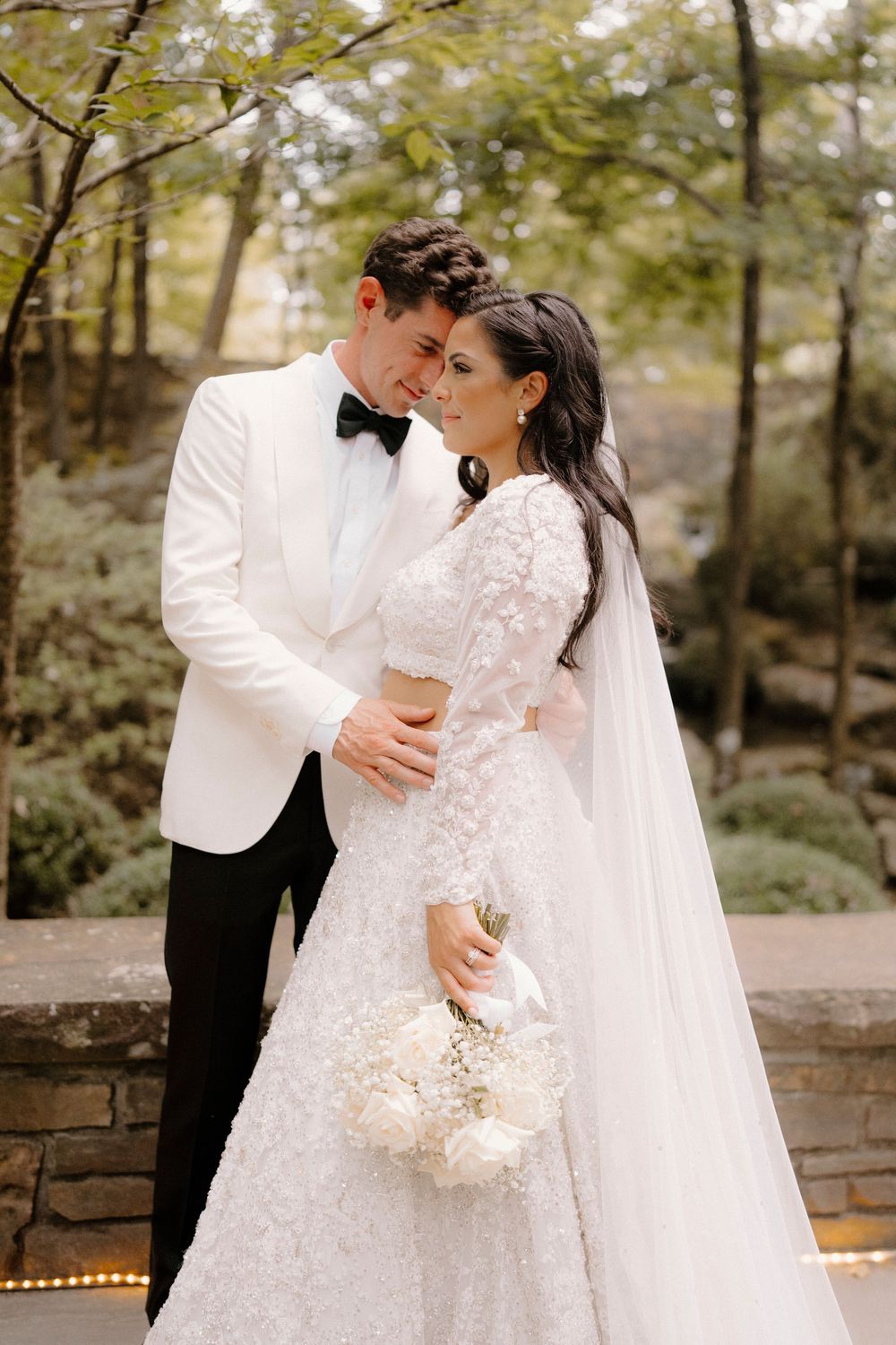 A romantic wedding couple embraces outdoors in a garden setting wearing elegant white attire and holding a bouquet.