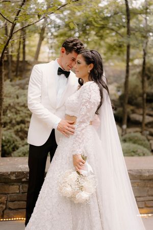 A romantic wedding couple embraces outdoors in a garden setting wearing elegant white attire and holding a bouquet.