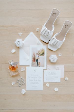 Wedding stationery flatlay with perfume bottle and white floral details arranged on light wooden surface.