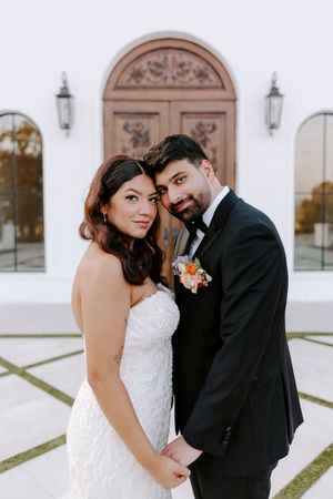 Newlywed couple poses together against a white church wall with elegant arched doorways and lanterns.