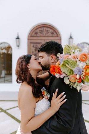Newlyweds embrace and kiss in front of ornate architecture while holding vibrant floral bouquet with orange blooms.