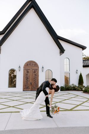 A romantic wedding couple dips in front of a white chapel with geometric patterned walkway and ornate wooden doors.