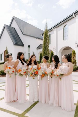 A wedding party in blush pink dresses stands in front of a modern white church building holding coral and peach bouquets.