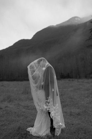 A flowing white veil blows dramatically in the wind against a misty mountain landscape backdrop.