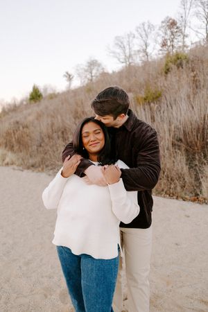 A couple embraces on a dirt path surrounded by winter trees and brush on a hillside.
