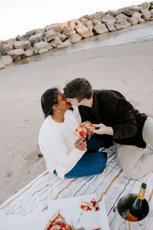 A couple shares a romantic picnic on a blanket by a rocky beach at sunset.