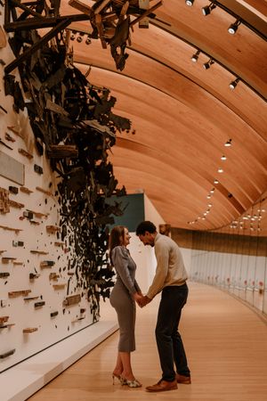 Two people share an intimate moment in a curved museum hallway with wooden ceiling and dramatic black wall installation.