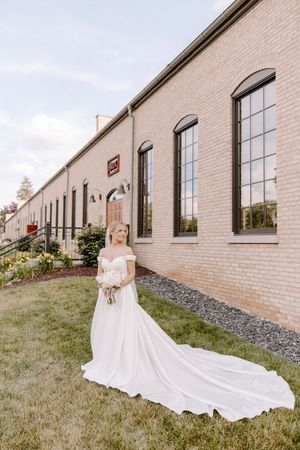 A bride in a flowing white off-shoulder wedding gown with long train stands beside a rustic brick building with arched windows.