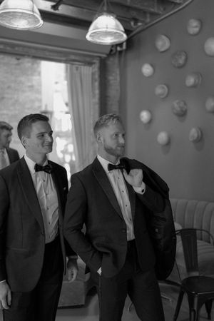 Four black and white photos show groomsmen in formal tuxedos laughing and smiling together at a wedding venue.