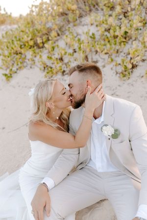 A couple in white attire shares romantic kisses while sitting on a sandy beach with foliage in the background.